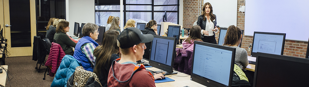 A group of students sit in a computer lab as a professor lectures.